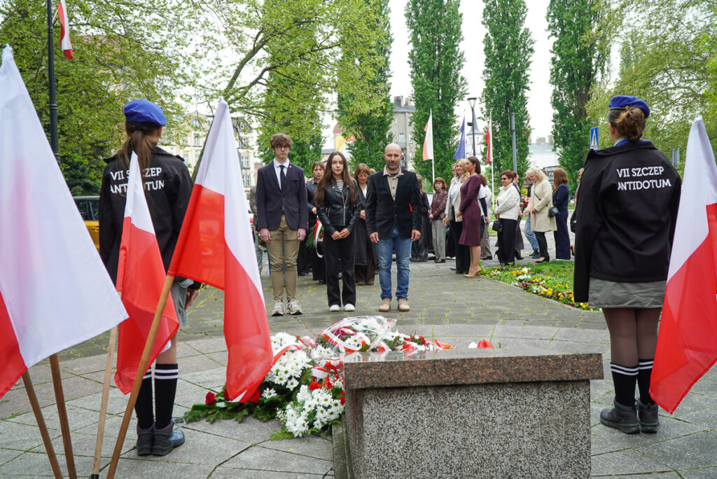 Delegacja mieszkańców i młodzieży stoi przed pomnikiem podczas obchodów Narodowego Dnia Zwycięstwa. Na pierwszym planie widoczne są wieńce i biało-czerwone kwiaty.