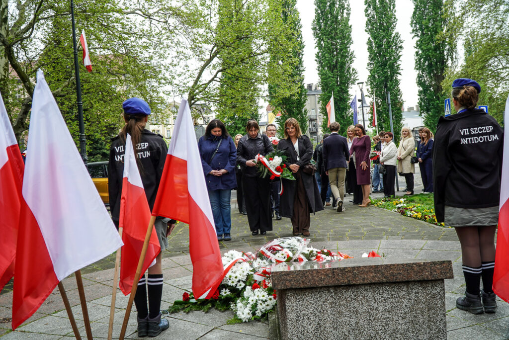 Grupa uczestników uroczystości podchodzi do pomnika z wiązanką kwiatów podczas obchodów Narodowego Dnia Zwycięstwa. W tle stoją mieszkańcy obserwujący ceremonię.