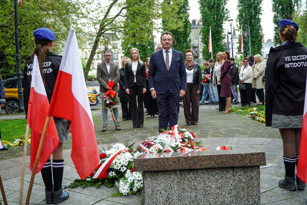 Delegacja uczestników uroczystości stoi przed pomnikiem pamięci podczas obchodów Narodowego Dnia Zwycięstwa. Wokół pomnika leżą biało-czerwone wieńce i kwiaty.