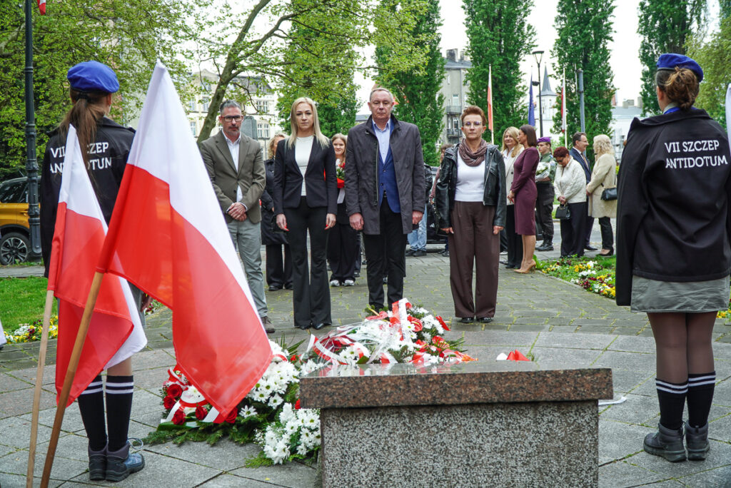 Uczestnicy uroczystości stoją przed pomnikiem podczas obchodów Narodowego Dnia Zwycięstwa. Na pierwszym planie widoczne są polskie flagi oraz liczne biało-czerwone wiązanki kwiatów złożone pod pomnikiem.