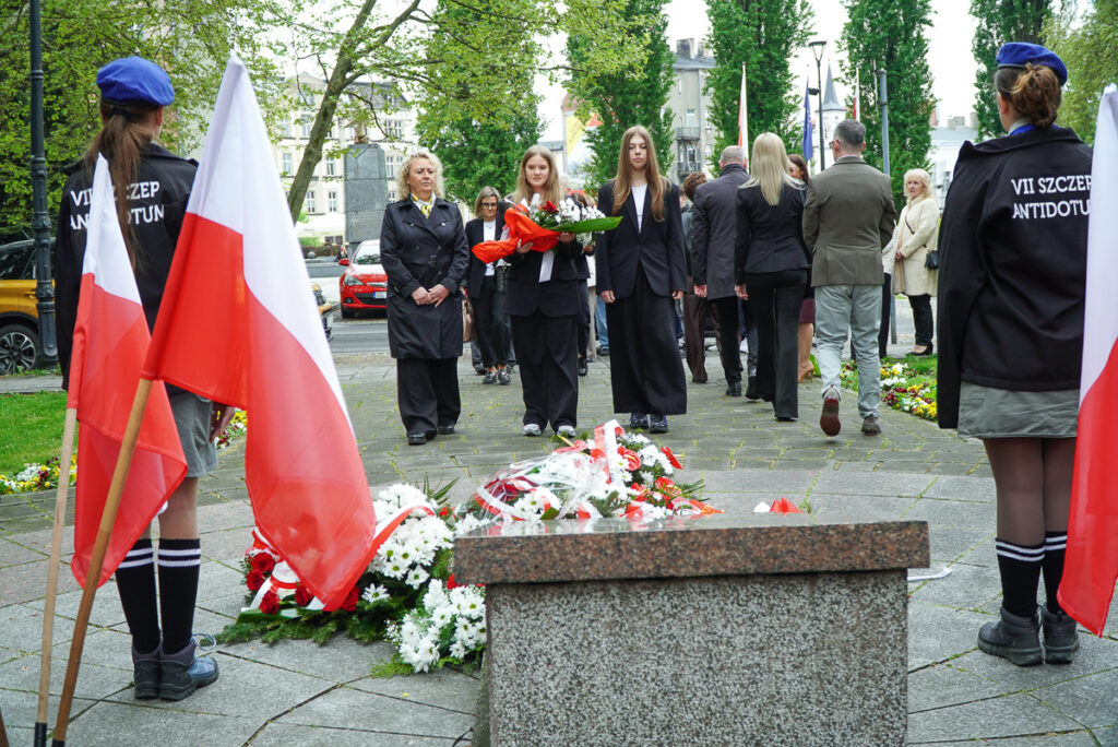 Dwie młode uczestniczki uroczystości niosą wiązankę biało-czerwonych kwiatów w stronę pomnika. Po obu stronach alejki stoją harcerze oraz ustawione są polskie flagi.