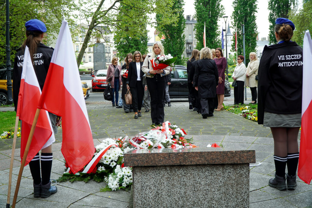 Uczestniczka uroczystości niesie biało-czerwoną wiązankę kwiatów w kierunku pomnika. W tle widoczni są pozostali uczestnicy wydarzenia oraz mieszkańcy obserwujący ceremonię.