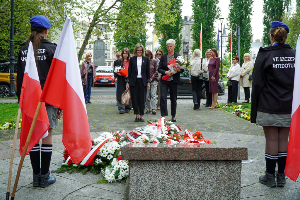 Dwie kobiety z wiązankami kwiatów podchodzą do pomnika podczas uroczystości Narodowego Dnia Zwycięstwa. Wokół pomnika leżą liczne biało-czerwone wieńce i kwiaty.
