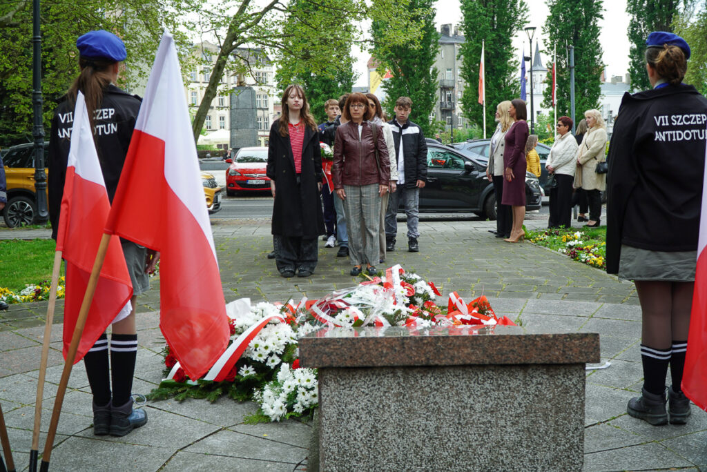 Grupa uczestników uroczystości stoi przed pomnikiem podczas chwili zadumy. Na pierwszym planie widoczne są wieńce i biało-czerwone kwiaty złożone pod pomnikiem.