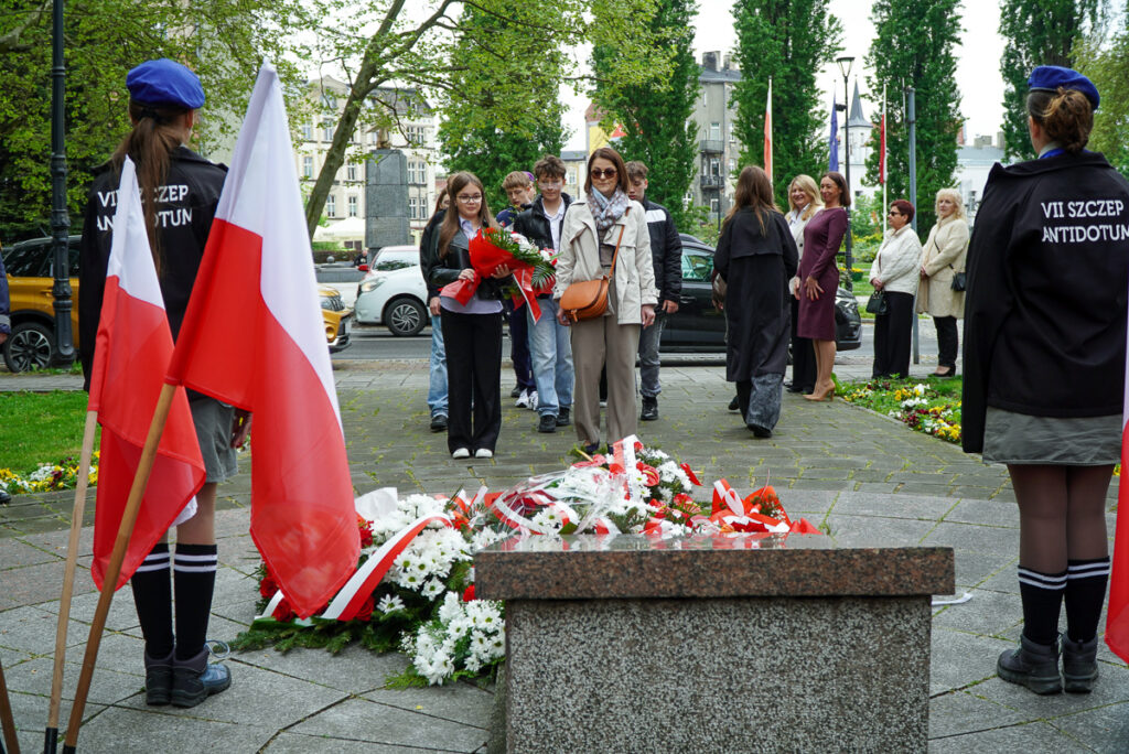 Młodzież i dorośli uczestnicy uroczystości podchodzą do pomnika z wiązanką biało-czerwonych kwiatów. Po bokach alejki stoją harcerze oraz polskie flagi.