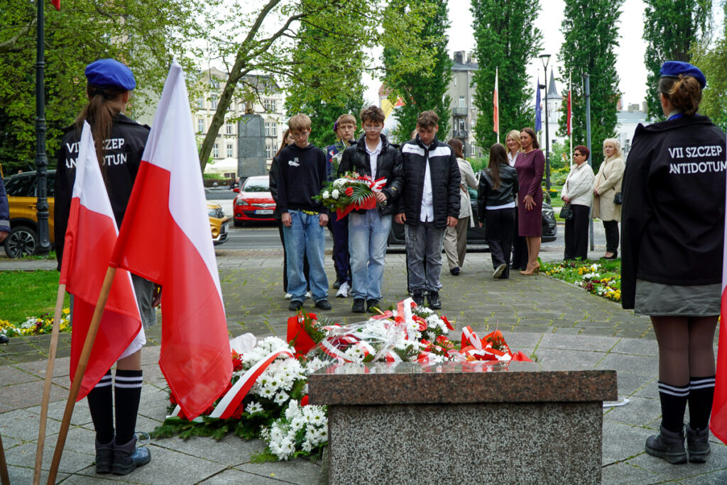Grupa młodych uczestników uroczystości niesie kwiaty w stronę pomnika podczas obchodów Narodowego Dnia Zwycięstwa. W tle stoją mieszkańcy obserwujący wydarzenie.