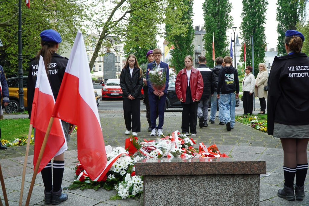 Trójka młodych uczestników uroczystości stoi przed pomnikiem z wiązanką kwiatów podczas obchodów Narodowego Dnia Zwycięstwa. Wokół pomnika znajdują się liczne biało-czerwone wieńce.