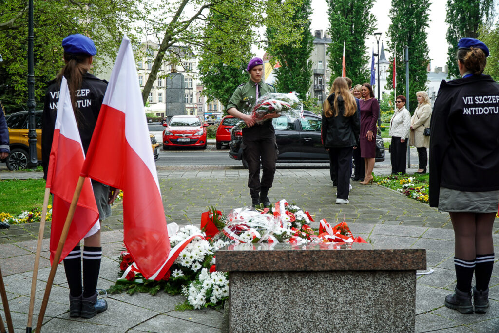 Harcerz w mundurze i berecie niesie dużą wiązankę kwiatów w kierunku pomnika podczas obchodów Narodowego Dnia Zwycięstwa. W tle widoczni są uczestnicy wydarzenia.