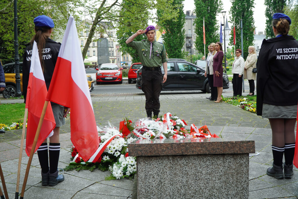 Harcerz stojący przed pomnikiem oddaje salut podczas obchodów Narodowego Dnia Zwycięstwa. Wokół pomnika znajdują się biało-czerwone wieńce i kwiaty, a po bokach stoją harcerze z flagami.