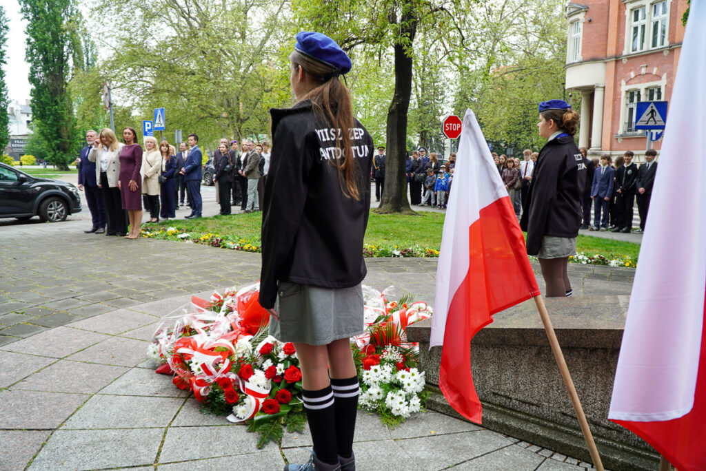 Widok na uczestników obchodów Narodowego Dnia Zwycięstwa zgromadzonych wokół pomnika pamięci. Na pierwszym planie stoją harcerze i polskie flagi, a przed pomnikiem leżą liczne biało-czerwone wieńce i kwiaty.