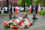 Dwie harcerki stoją na warcie honorowej przy pomniku pamięci. Przed pomnikiem leżą liczne biało-czerwone wieńce i kwiaty, a wokół ustawione są polskie flagi.