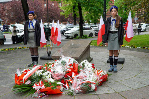 Dwie harcerki stoją na warcie honorowej przy pomniku pamięci. Przed pomnikiem leżą liczne biało-czerwone wieńce i kwiaty, a wokół ustawione są polskie flagi.