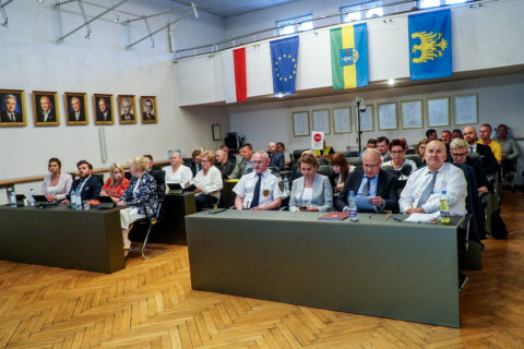 Widok na salę obrad wypełnioną radnymi, urzędnikami i gośćmi podczas 31. sesji Rady Miasta Siemianowic Śląskich. Nad uczestnikami wiszą flagi Polski, Unii Europejskiej, miasta oraz województwa śląskiego. W tle widoczne są portrety i tablice na ścianie sali.