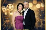 Plakat promujący koncert zatytułowany „Filmowy zawrót głowy", który odbędzie się w SCK- Bytków. Grafika utrzymana jest w eleganckiej, kinowej stylistyce, z dominującymi złotymi i ciemnymi odcieniami oraz motywami nawiązującymi do filmu i muzyki. W centralnej części plakatu znajduje się para artystów stojących obok siebie. Kobieta ubrana jest w elegancką, fioletową suknię bez ramiączek oraz naszyjnik, a mężczyzna w ciemny garnitur i jasną koszulę. Oboje patrzą w stronę odbiorcy, stojąc blisko siebie w spokojnej, formalnej pozie. Tło przedstawia stylizowaną scenę z rozchylonymi teatralnymi kurtynami, reflektorami oraz elementami związanymi z kinem, takimi jak taśmy filmowe i projektor. Widoczne są także graficzne motywy nut i pięciolinii, podkreślające muzyczny charakter wydarzenia.