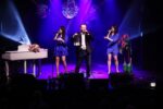 Na scenie SCK – Bytków występują Damian Holecki oraz dwie wokalistki trzymające mikrofony. Artyści śpiewają w kolorowym, fioletowo-niebieskim oświetleniu scenicznym. Po lewej stronie widoczny jest biały fortepian udekorowany kwiatami, a scena ozdobiona jest kompozycjami florystycznymi i lampionami.