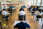 Sala gimnastyczna przystosowana do egzaminu maturalnego – w równych rzędach ustawione są pojedyncze stoliki, przy których siedzą uczniowie w odświętnych strojach, skupieni na pisaniu arkusza. Na pierwszym planie widać uczennicę siedzącą tyłem, z arkuszem egzaminacyjnym i długopisem na ławce. W tle, przy stole komisji egzaminacyjnej, siedzą nauczyciele nadzorujący przebieg egzaminu. Na ścianie widoczny jest zegar oraz polskie flagi. W lewym górnym rogu grafiki znajduje się napis „Matura 2026”, a w dolnym logo Siemianowic Śląskich.