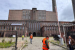 Ludzie w pomarańczowych kamizelkach stoją na wybrukowanej "kocimi łbami" ulicy. W tle monumentalna budowla industrialna.