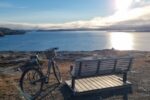 Fotografia przedstawia rower stojący obok ławki na skalistym brzegu nad spokojną zatoką, oświetloną niskim słońcem odbijającym się w wodzie.