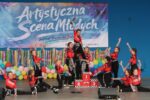 Fotografia przedstawia występ dziecięcego zespołu tanecznego Ti-Top podczas wydarzenia „Artystyczna Scena Młodych”. Młodzi artyści ubrani są w sportowo-sceniczne stroje w kolorach czerwieni, czerni i bieli, ozdobione motywem szachownicy. Tancerze wykonują efektowną choreografię, tworząc wielopoziomową kompozycję sceniczną z centralnie ustawionym podium. Niektóre dzieci pozują z uniesionymi rękami, inne wykonują szpagaty lub wspierają główną postać znajdującą się najwyżej w układzie. Kolorowe balony i dekoracyjne wstążki w tle podkreślają radosny, festiwalowy charakter wydarzenia. Zdjęcie oddaje energię występu, zaangażowanie młodych wykonawców oraz atmosferę wspólnej scenicznej pasji.