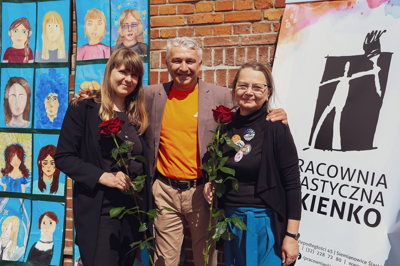 Trzy osoby pozują do zdjęcia przy plenerowej wystawie dziecięcych autoportretów. Obok znajduje się baner Pracowni Plastycznej „Okienko”.