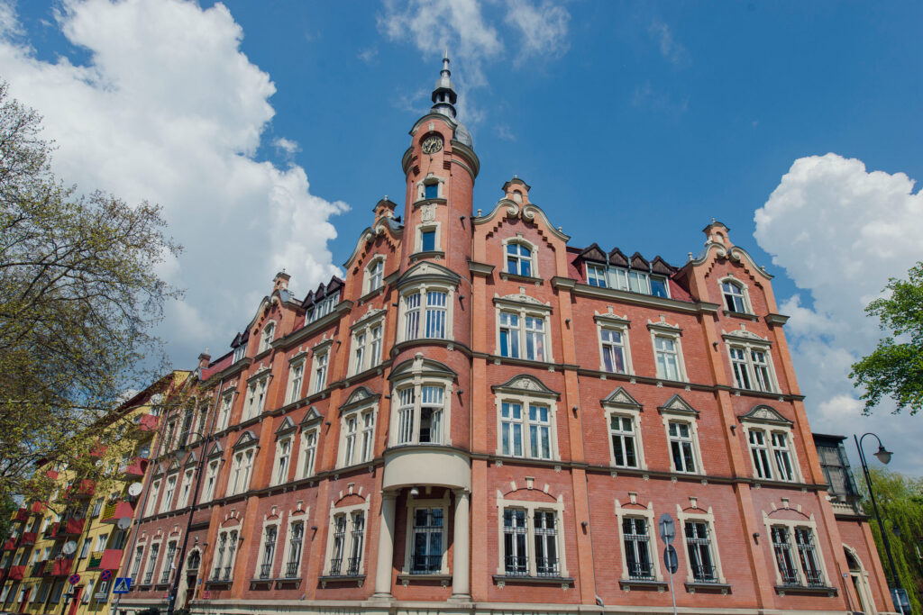 Zdjęcie przedstawia zabytkowy, wielopiętrowy budynek z czerwonej cegły, usytuowany na rogu ulicy. Jego najbardziej charakterystycznym elementem jest zaokrąglony narożnik zwieńczony ozdobną wieżą zegarową ze spiczastym hełmem. Fasada budynku charakteryzuje się licznymi, rytmicznie rozmieszczonymi oknami z jasnymi, ozdobnymi obramowaniami. Na parterze znajdują się rzędy okien z metalowymi kratami. Powyżej widać zdobione wykusze i gzymsy. Budynek otoczony jest drzewami z wiosennymi liśćmi po lewej stronie oraz fragmentem zieleni po prawej. W tle, po lewej stronie, widoczny jest blok mieszkalny o żółtej fasadzie. Niebo jest błękitne z białymi, puszystymi chmurami. Na pierwszym planie, przy krawężniku, widać znaki drogowe.