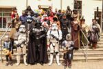 Grupa osób w kostiumach inspirowanych uniwersum Star Wars pozuje do wspólnego zdjęcia na schodach przed budynkiem. Na pierwszym planie stoją m.in. Darth Vader, szturmowcy oraz dziecko przebrane za Mandalorianina.