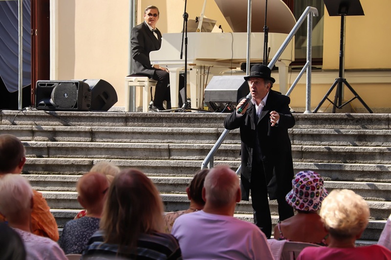 Dwóch artystów występuje na schodach przed budynkiem. Wokalista śpiewa do publiczności, a pianista akompaniuje na białym fortepianie. Zdjęcie promujące koncert z cyklu „Muzyka u Rheinbabenów”.