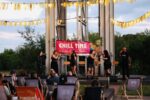 Zdjęcie przedstawia letni koncert plenerowy z cyklu „Muzyczne Lato. CHILL TIME pod szybem Krystyn”. Na podwyższeniu zadaszonym konstrukcją szybu występuje sześcioosobowy zespół wokalny (cztery kobiety i dwóch mężczyzn). Wszyscy artyści są ubrani w czarne stroje, trzymają mikrofony i śpiewają, poruszając się na scenie. W tle, za plecami artystów, rozciągnięty jest duży, różowy baner z białym napisem informującym o nazwie wydarzenia. Za banerem stoją puste drewniane krzesła barowe na wysokich nogach. Scena jest wkomponowana w monumentalną, jasnoszarą, metalową konstrukcję zabytkowego szybu kopalnianego, która dominuje w tle kadru. Po bokach widoczna jest zielona roślinność i drzewa. Na pierwszym planie, na trawiastym podłożu przed sceną, tyłem do obiektywu siedzą uczestnicy koncertu. Odpoczywają na rozstawionych leżakach, z których część ma szare oparcia z widocznym białym logo „SCK”.