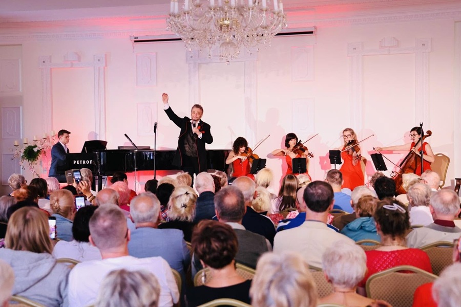 Koncert kameralny w eleganckiej sali z cyklu „Muzyka u Rheinbabenów”. Na scenie występuje wokalista, pianista przy czarnym fortepianie oraz cztery instrumentalistki w czerwonych sukienkach, a występowi przygląda się liczna publiczność. Scena i ściany sali są białe, ozdobione klasycznymi sztukateriami oraz ozdobnymi obramowaniami. U sufitu, w górnej centralnej części kadru, wisi duży, kryształowy, wieloramienny żyrandol. Dolną połowę kadru zajmują odwróceni tyłem do obiektywu widzowie. Publiczność, w której przeważają starsze osoby, siedzi blisko siebie na krzesłach i w skupieniu ogląda występ/