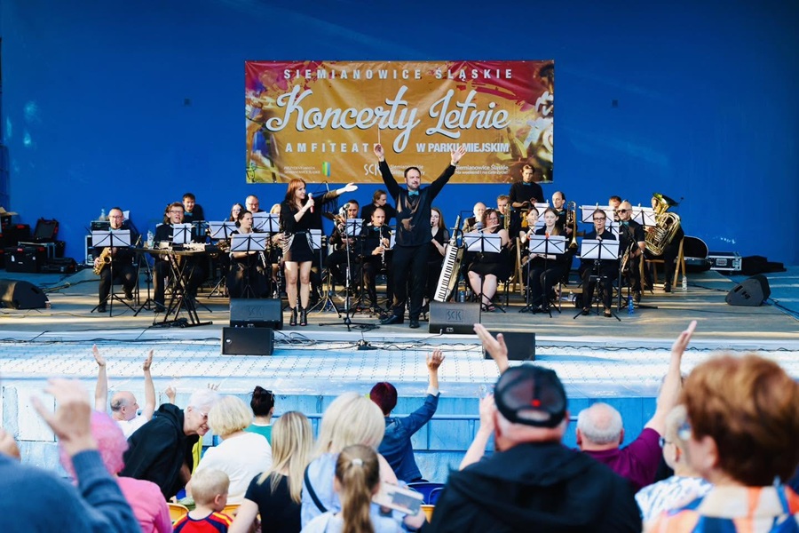 Zdjęcie przedstawia żywiołowy koncert plenerowy. Na dużej scenie amfiteatru występuje orkiestra dęta, składająca się z kilkunastu muzyków (kobiet i mężczyzn) ubranych w eleganckie, ciemne stroje, siedzących przy pulpitach z nutami. Widoczne są instrumenty takie jak saksofony, trąbki, klarnety oraz puzony i tuba. Dolną część kadru zajmuje widownia uwieczniona od tyłu. Zgromadzeni ludzie w różnym wieku siedzą na amfiteatralnych trybunach. Wiele osób ma uniesione w górę ręce – klaszczą, machają lub filmują występ telefonami, co podkreśla radosną i energiczną atmosferę koncertu.
