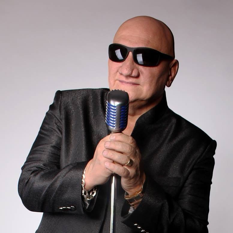 Mężczyzna w ciemnym stroju i okularach przeciwsłonecznych trzyma mikrofon. Zdjęcie promujące koncert Piotra Herdziny podczas letnich koncertów w amfiteatrze w Siemianowicach Śląskich.