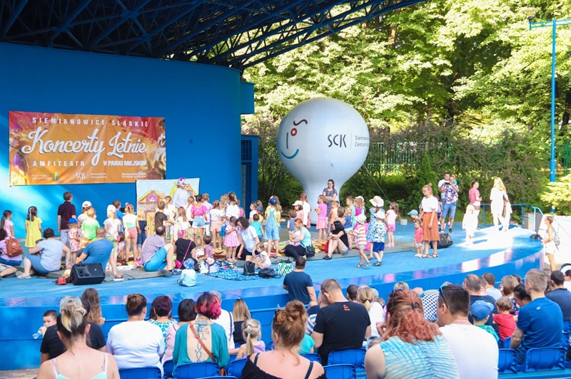 Dzieci wraz z opiekunami uczestniczą w spektaklu plenerowym na scenie amfiteatru w Parku Miejskim w Siemianowicach Śląskich. Na scenie trwa interaktywne przedstawienie dla najmłodszych.