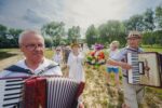 Senioralna Noc Świętojańska na Rzęsie. Mężczyzna z akordeonem prowadzi korowód w którym idą przebrani uczestnicy z wiankami na głowie.