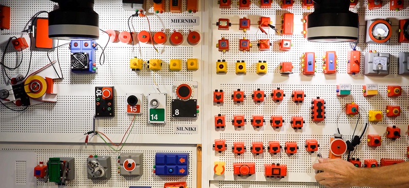 Tablica laboratoryjna z zamontowanymi modułami elektronicznymi i elektrycznymi. Widoczne są wskaźniki, przełączniki, silniki oraz ręka osoby sięgającej po jeden z elementów.