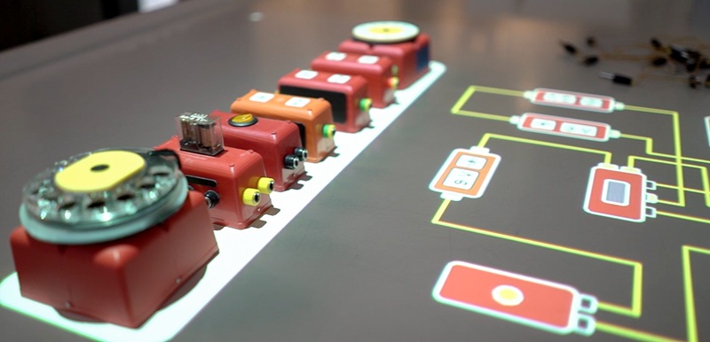 Rząd czerwonych i pomarańczowych modułów elektrotechnicznych ustawionych na stole obok schematu obwodu elektrycznego.