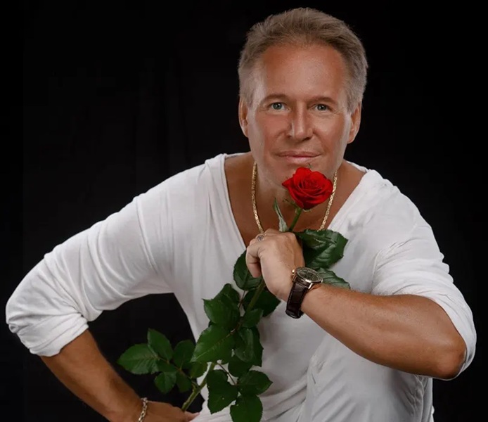 Portret Jacka Silskiego na czarnym tle. Mężczyzna ubrany w białą bluzkę z długim rękawem patrzy z uśmiechem w stronę obiektywu, trzymając w dłoni czerwoną różę z zielonymi liśćmi. Na szyi ma złoty łańcuszek, a na nadgarstku zegarek na ciemnym pasku.
