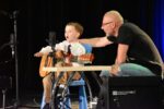 Mały chłopiec w białej koszuli i krawacie siedzi na niebieskim krzesełku na scenie, gra na małej gitarze klasycznej i śpiewa do mikrofonu. Obok niego przykuca mężczyzna w czarnej koszulce i okularach, trzymając dłoń wspierająco na ramieniu chłopca. W tle widać czarną kurtynę sceniczną.