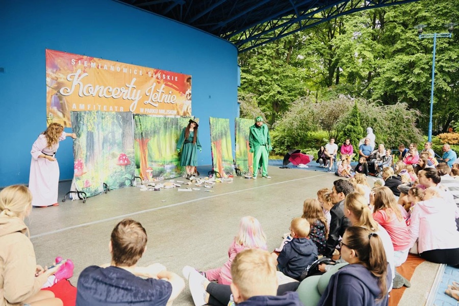 Spektakl teatralny dla dzieci w amfiteatrze miejskim. Na scenie z dekoracjami przedstawiającymi bajkowy las występują aktorzy w przebraniach: kobieta w różowej sukni oraz dwie postacie w zielonych strojach, w tym jedna w przebraniu żaby. Przed sceną siedzą na ziemi zgromadzone dzieci wraz z opiekunami i oglądają przedstawienie. W tle widać niebieską ścianę sceny z banerem „Koncerty Letnie” oraz parkowe drzewa.