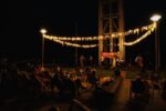 Nocny koncert plenerowy z cyklu „Chill Time pod szybem Krystyn”. Zespół muzyczny występuje na podwyższeniu pod oświetloną wieżą szybu wyciągowego. Przed sceną publiczność wypoczywa na leżakach. Przestrzeń rozświetlają ciepłym światłem latarnie oraz zawieszone między nimi girlandy świetlne z dekoracyjnymi wstążkami. Panuje kameralny, wieczorny nastrój.