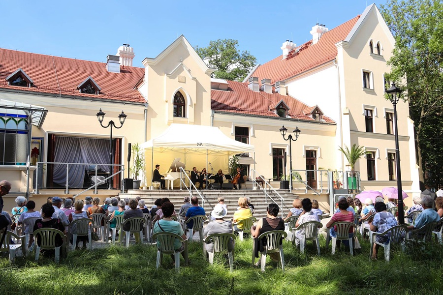 Plenerowy koncert muzyki klasycznej przed zabytkowym, jasnym zameczkiem z czerwonym dachem. Zespół muzyczny (pianista przy białym fortepianie oraz instrumentaliści smyczkowi) występuje w białym namiocie na tarasie budynku. Przed schodami, na trawie, na białych krzesłach ogrodowych siedzą widzowie słuchający koncertu w słoneczny dzień.