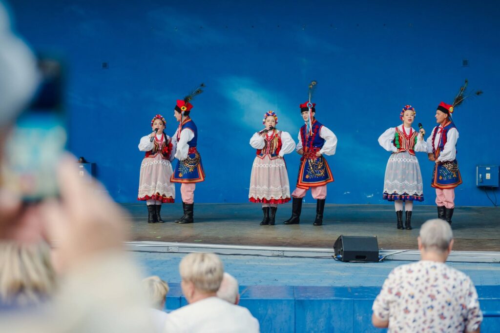 Zespół folklorystyczny w tradycyjnych krakowskich strojach ludowych (czerwone gorsety, białe koszule, spódnice w kwiaty, czapki z pawimi piórami) śpiewa do mikrofonów na scenie z niebieskim tłem, przed publicznością obserwującą występ z widowni.