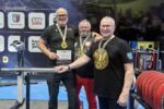 Trzech mężczyzn w czarnych koszulkach sportowych z napisami i logotypami pozuje do wspólnego zdjęcia podczas Mistrzostw Świata Federacji GPC w sportach siłowych. Wszyscy mają na szyjach medale, a dwóch z nich trzyma dyplomy. W tle znajduje się baner z logotypami organizatorów i sponsorów oraz nazwami partnerów wydarzenia. Po lewej stronie widoczna jest flaga Polski, a przed zawodnikami stoją stojaki ze sztangą.