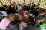 Grupa uczniów zgromadzona wokół dużego drewnianego stołu w altanie. Młodzież przygląda się materiałom edukacyjnym i przyrządom laboratoryjnym przygotowanym do zajęć.