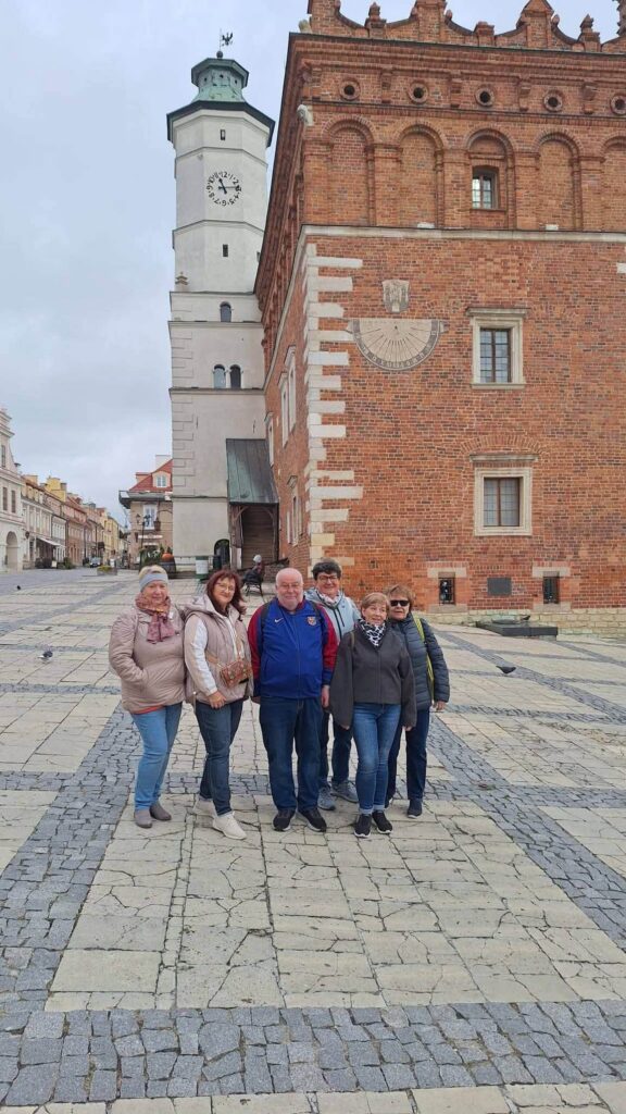 Sześcioosobowa grupa turystów pozuje na sandomierskim rynku. W tle zabytkowy ceglany ratusz z zegarem słonecznym oraz wysoka wieża zegarowa.